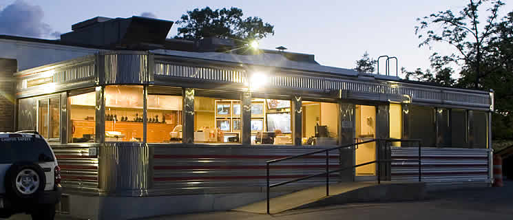 A vintage-style diner with large windows and metal siding illuminated at dusk. The interior is warmly lit, and a ramp leads up to the entrance. Trees and a parked vehicle are visible in the background.